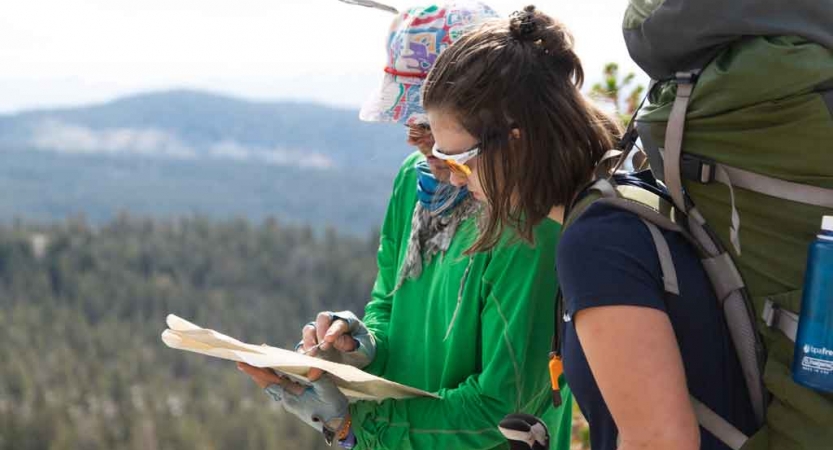 Two people wearing backpacks examine a map. There is a vast landscape in the background with evergreen trees and mountains